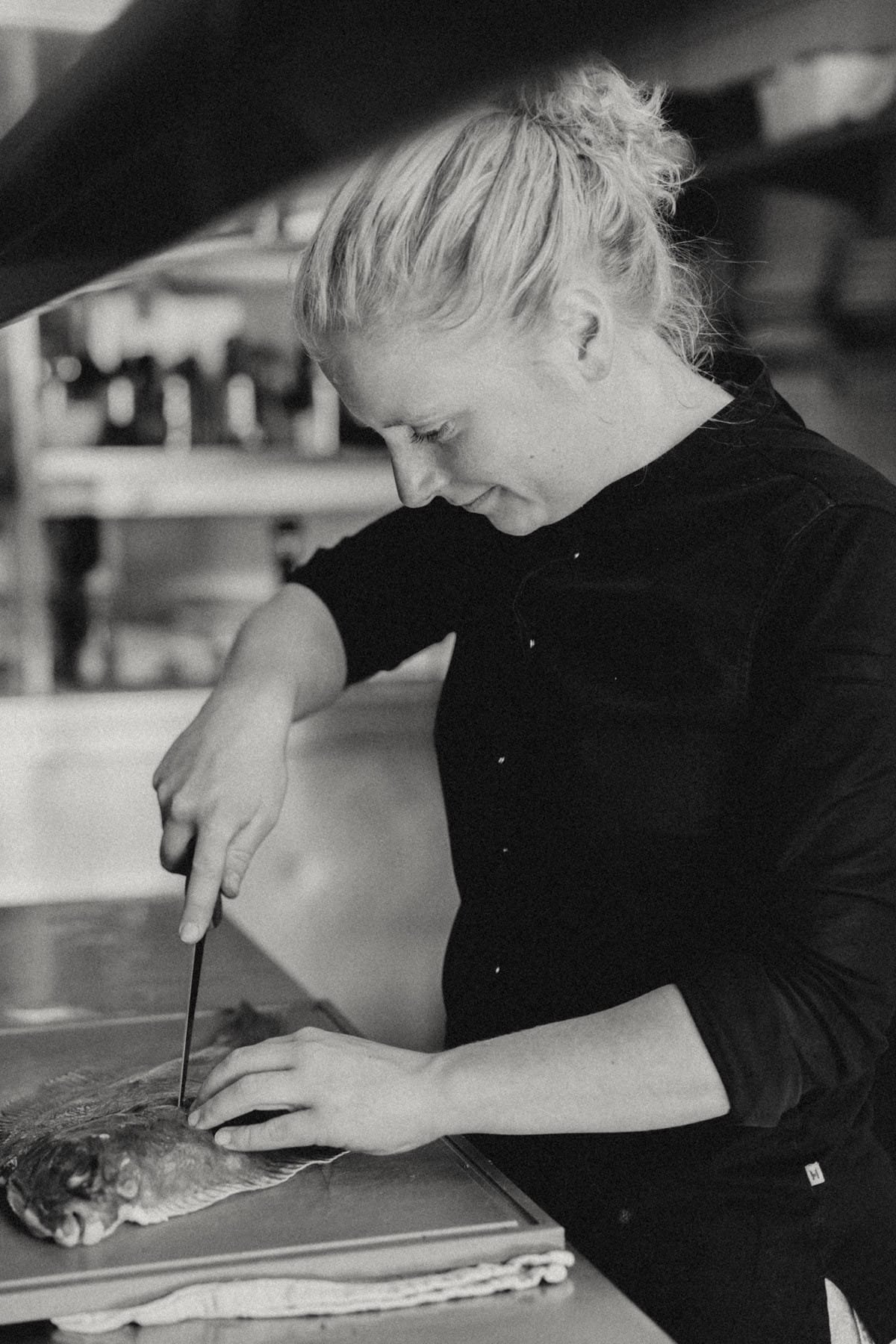 Een chef in een zwarte schort fileert een vis met een mes op een werkblad in de keuken van een restaurant.